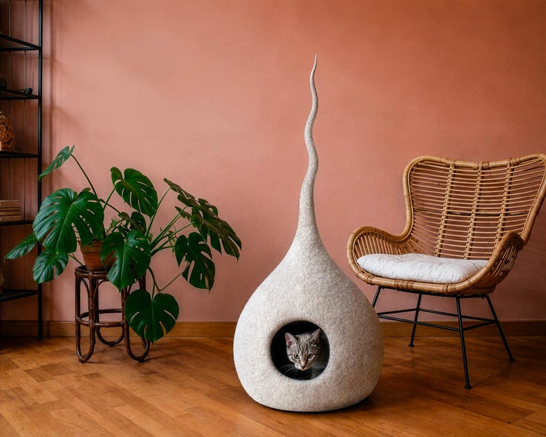 K&ouml;nnte beinhalten: Ein modernes Katzenbett in hellem Grau mit einer hohen, geschwungenen Form. Das Bett hat eine runde &Ouml;ffnung, in der eine graue Tabby-Katze zu sehen ist. Ein Topf mit einer Pflanze und ein Korbstuhl sind im Hintergrund.