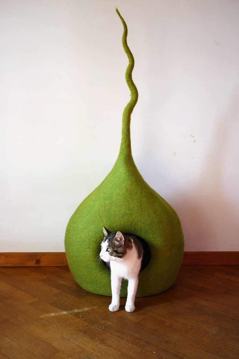 May include: A green felt cat bed shaped like a teardrop with a long, curved stem. A cat is peeking out of the opening of the bed.
