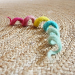 Puede incluir: Una cuerda de fieltro rizada de colores rosa, amarillo y azul sobre una superficie tejida de color marr&oacute;n.