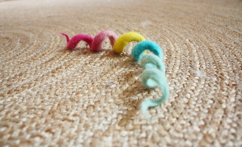 Puede incluir: Una cuerda de fieltro rizada de colores rosa, amarillo y azul sobre una superficie tejida de color marr&oacute;n.