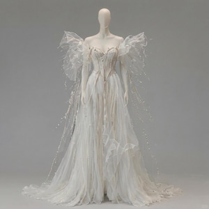 Può includere: Un abito bianco lungo fino ai piedi caratterizzato da un corpetto a corsetto strutturato con decorazioni di perle verticali, maniche in tulle arricciate scoperte sulle spalle e una gonna fluida. Il capo è ornato da lunghi e delicati fili di cristalli.