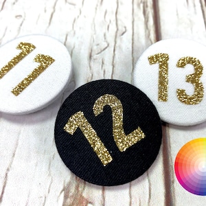 May include: Three round buttons with gold glitter numbers on a white wood background. The numbers are 11, 12, and 13. The button with the number 12 is black.