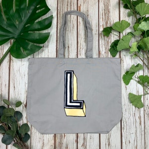 May include: A gray tote bag with a large, stylized letter "L" in black, white, and yellow. The tote bag has gray handles and is made of a soft fabric. The design is a bold, graphic statement.