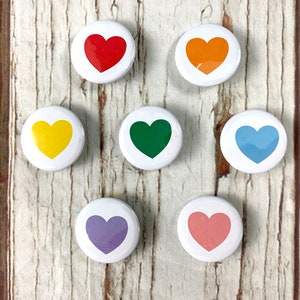 May include: Seven round white buttons with a heart design in different colours. The colours are red, orange, yellow, green, blue, purple, and pink.