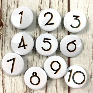 Może przedstawiać: Dziesięć białych okrągłych guzików z czarnymi cyframi od 1 do 10. Cyfry mają cienki pomarańczowy kontur.