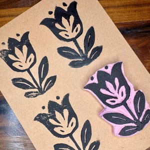 May include: A light pink rubber stamp featuring a stylized tulip design with black ink applied to the raised surface. The stamp is positioned next to three black ink impressions of the same tulip motif printed on a rectangular piece of tan paper.