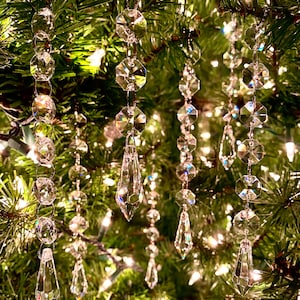 Op de afbeelding: Kristallen strengen met traanvormige hangers die aan een groene kerstboom hangen. De strengen zijn gemaakt van heldere glazen kralen en zijn aan elkaar geregen met zilverdraad.
