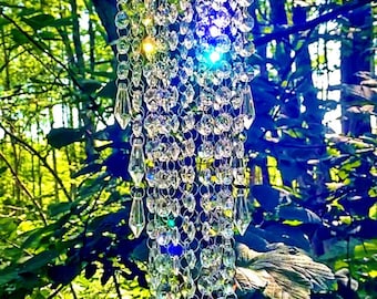 May include: A crystal suncatcher hanging from a tree branch in a forest. The suncatcher is made of many small crystals that sparkle in the sunlight. The crystals are arranged in a cascading pattern, creating a beautiful and whimsical effect.