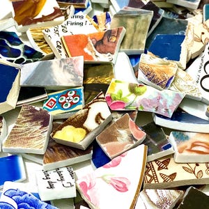 May include: A close-up of a pile of broken ceramic tiles, showcasing a variety of patterns and colours. The fragments feature floral designs, abstract art, and text, with hues of blue, pink, brown, and white. Some pieces have visible text.