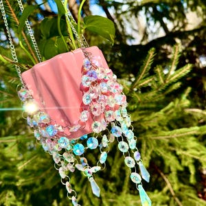 May include: A pink ceramic planter with a green plant hanging from a silver chain. The planter is decorated with iridescent crystal beads and teardrop-shaped crystals.