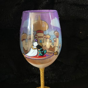 May include: A clear glass wine glass with a gold glitter stem and base. The glass is decorated with a hand-painted scene of Aladdin and Jasmine on a magic carpet flying over the palace in Agrabah.