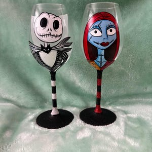 May include: Two hand-painted wine glasses featuring characters from The Nightmare Before Christmas. The glass on the left features Jack Skellington, the Pumpkin King, in black and white. The glass on the right features Sally, Jack's ragdoll girlfriend, in red, blue, and white.