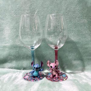 May include: Two clear wine glasses with glitter stems, one blue and one pink. Each glass features a character figurine at the base, surrounded by colorful rhinestones. The figurines are a blue Stitch and a pink Angel.