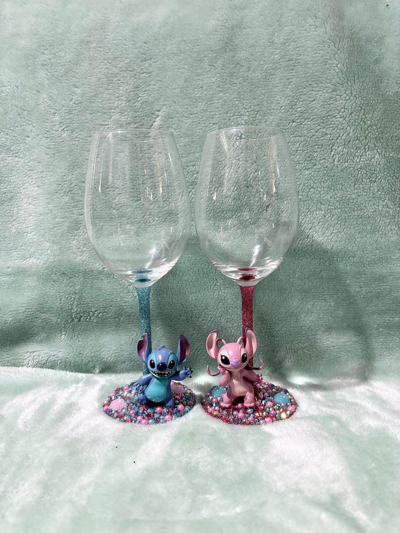 May include: Two clear wine glasses with glitter stems, one blue and one pink. Each glass features a character figurine at the base, surrounded by colorful rhinestones. The figurines are a blue Stitch and a pink Angel.