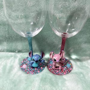 May include: Two clear wine glasses with glitter stems, one blue and one pink. Each glass has a character figurine at the base, one blue and one pink, surrounded by colorful gems. The glasses are on a light blue surface.