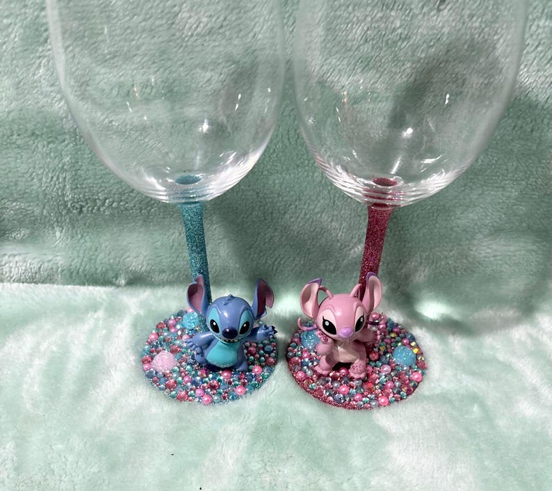May include: Two clear wine glasses with glitter stems, one blue and one pink. Each glass has a character figurine at the base, one blue and one pink, surrounded by colorful gems. The glasses are on a light blue surface.