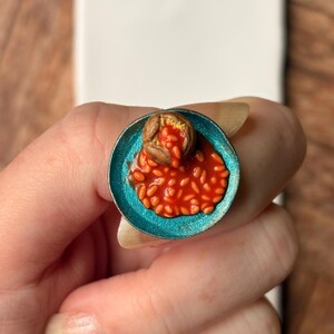 May include: A miniature ring featuring a small teal-coloured plate topped with a brown baked potato and a generous serving of orange baked beans in tomato sauce. The food items are textured and realistic, set within a circular metal plate base.