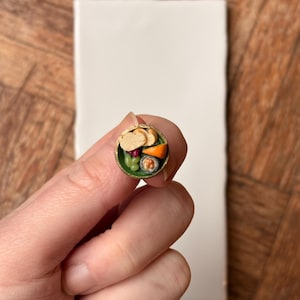 Puede incluir: Un pequeño cuenco verde con borde dorado, lleno de mini sushi y guarniciones. El cuenco contiene rollos de sushi, pan y verduras. El cuenco está en la mano de una persona.