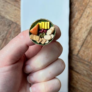 Puede incluir: Mini tabla de embutidos redonda con galletas, queso, uvas y embutidos. La tabla tiene un borde verde y se sostiene en una mano. El fondo muestra una baldosa blanca y una superficie de madera.