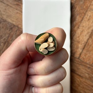 May include: A miniature food ring with a baguette and sliced bread pieces, set against a green background within a gold-coloured ring. The ring is held by a hand, with a white rectangular tile and wooden surface in the background.
