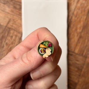 Puede incluir: Plato de comida en miniatura con borde verde, lleno de pequeños sushi, verduras y un elemento en forma de pan. El plato se sostiene en la mano de una persona, con una superficie rectangular blanca y un fondo de madera.