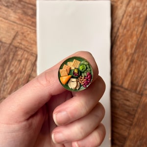 Puede incluir: Un dije miniatura, redondo, con borde verde, que representa una detallada tabla de charcutería. La tabla incluye galletas saladas, queso, aceitunas, frijoles y otros alimentos pequeños. El dije se sostiene en una mano contra una baldosa blanca y una superficie de madera.