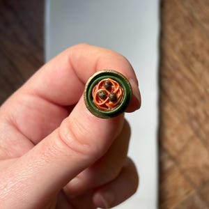 May include: A miniature, handcrafted ring featuring a detailed spaghetti and meatballs design. The ring has a green and gold border, with orange spaghetti and three brown meatballs. The ring is held against a white and brown background.
