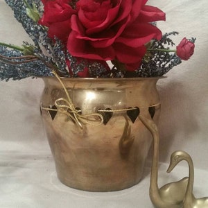May include: A brass planter with a heart-shaped cutout design around the rim. The planter is filled with a bouquet of red roses and purple flowers. Two gold swan figurines are in the foreground.