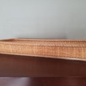 May include: A rectangular woven basket in a light brown color. The basket has a flat base and a slightly raised rim. The weave is tight and even, creating a sturdy container. The basket is empty and sits on a dark brown surface.