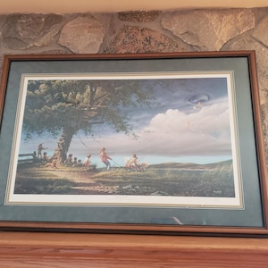 May include: Framed print depicting a serene outdoor scene with figures near a large tree. The artwork features a blue sky with clouds, a grassy area, and a path. The frame is brown with a green mat.