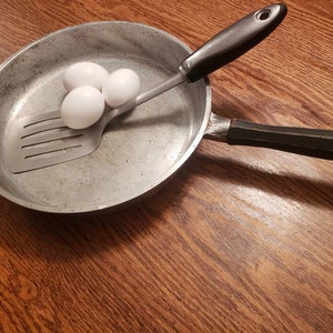 May include: A silver frying pan with a black handle and a silver spatula inside. Three white eggs are sitting on the spatula.