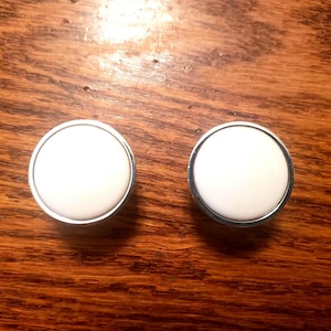 May include: Two round cabinet knobs with a white center and a silver-colored metal rim. The knobs are set against a dark wood grain background. These knobs are suitable for furniture or home decor projects.