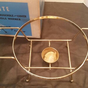 K&ouml;nnte beinhalten: Goldfarbener Kerzenw&auml;rmer aus Metall mit einem wei&szlig;en Keramikgriff. Der W&auml;rmer hat eine runde Basis und eine zentrale Vertiefung f&uuml;r eine Kerze. Der W&auml;rmer befindet sich in seiner Originalverpackung, auf der "Anchorwhite Candle King 1 1/4 qt. Casserole and Cover Candle Warmer" steht.