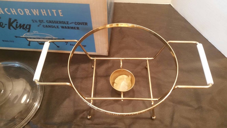 K&ouml;nnte beinhalten: Goldfarbener Kerzenw&auml;rmer aus Metall mit einem wei&szlig;en Keramikgriff. Der W&auml;rmer hat eine runde Basis und eine zentrale Vertiefung f&uuml;r eine Kerze. Der W&auml;rmer befindet sich in seiner Originalverpackung, auf der "Anchorwhite Candle King 1 1/4 qt. Casserole and Cover Candle Warmer" steht.