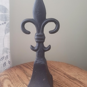 May include: A black fleur-de-lis sculpture, standing upright on a wooden surface. The sculpture has a pointed top, curved side elements, and a textured finish. The design is a classic symbol of royalty and heraldry.