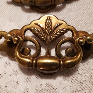 May include: Close-up of a brass drawer pull with a wheat stalk design. The pull has a curved handle and decorative details. The hardware is a warm gold color and appears to be vintage.