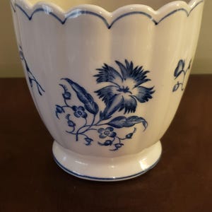 May include: White ceramic planter with a scalloped rim and a pedestal base. The planter is decorated with blue floral patterns, including a large flower and smaller sprigs. The rim is also trimmed with blue.