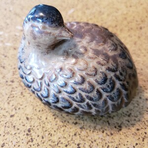 May include: A ceramic duck figurine with a speckled, textured body in shades of brown, beige, and blue. The duck has a dark blue head and a small, pointed beak. The figurine is a decorative item.