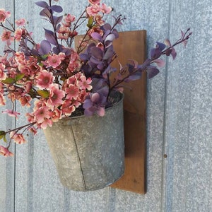 May include: A metal bucket planter with pink and purple flowers hanging on a wooden board attached to a gray wall.
