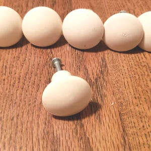 May include: Cream-colored, round cabinet knobs with a single knob in the foreground. The knobs are arranged on a wooden surface. The knobs have a smooth, matte finish and are likely made of ceramic or a similar material.