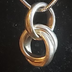 May include: Silver-toned pendant necklace featuring an interlocking rings design. The pendant has a polished finish, with two rings intertwined. The necklace chain is a delicate, silver-colored snake chain. The background is dark.
