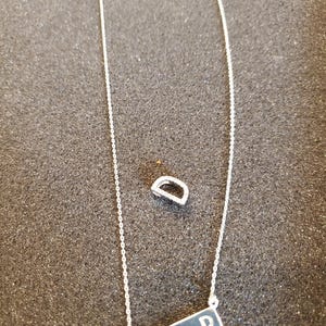 May include: A silver necklace with a rectangular pendant and the letter "D" engraved on it. A matching silver ring is also present. The necklace chain is delicate, and the pendant has a dark background.