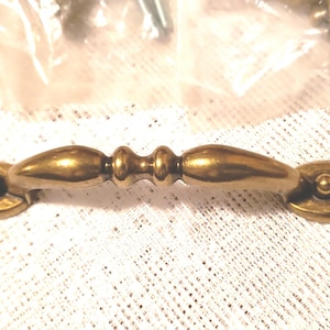 May include: A close-up of a brass-colored cabinet pull. The pull has an oval base with a decorative design and a curved handle. The hardware is new and appears to be unused. The background is a white, textured surface.
