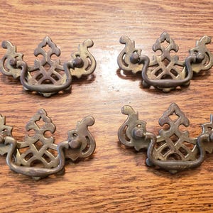 May include: Set of six antique brass drawer pulls. Each pull features an ornate, openwork design with a central handle and decorative side elements. The pulls are arranged on a wooden surface, showcasing their aged patina and vintage aesthetic.