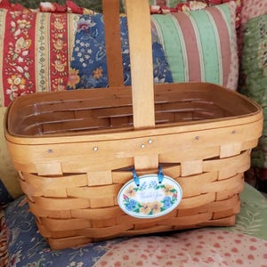 May include: A light brown woven basket with a wooden handle. The basket has a rectangular shape and a decorative tag that says "Thank You" with floral accents. The basket is sitting on a floral patterned pillow.
