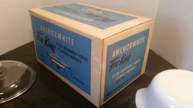 K&ouml;nnte beinhalten: Eine antike Anchorwhite Fire-King 1,5 Liter Auflaufform mit Deckel und einer blauen und wei&szlig;en Schachtel. Die Schachtel tr&auml;gt den Text "Anchorwhite Fire-King 1 1/2 qt. Casserole and Cover Candle Warmer".