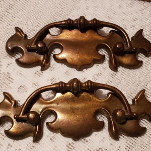 May include: Two antique bronze-colored drawer pulls. Each pull has a decorative, curved handle and a central, ornate plate. The pulls are made of metal and have a vintage aesthetic. They are designed for furniture restoration or decorative purposes.