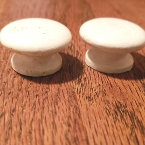 May include: Two white, round cabinet knobs with a mushroom-shaped design. The knobs are made of a solid material and sit on a wooden surface. The knobs are a light cream color and have a simple, classic design.
