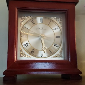 May include: A rectangular, wooden Seth Thomas clock with a burgundy finish. The clock face is gold with black Roman numerals and ornate gold detailing. The clock has a glass front and sits on a wooden base.