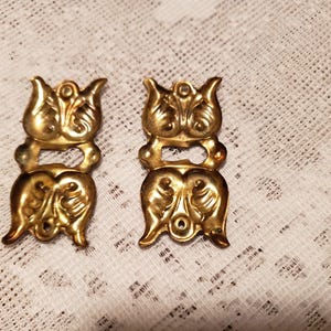 May include: Two ornate, gold-colored metal clasps. Each clasp features a decorative, symmetrical design with floral and scroll-like elements. The clasps are likely for decorative purposes or to secure a piece of furniture or jewelry.